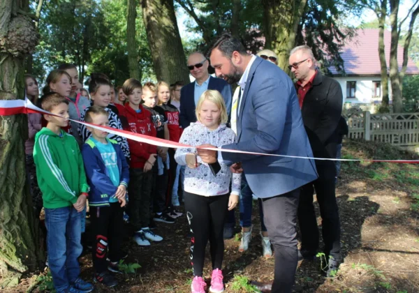 Grupa dzieci i dorosłych podczas uroczystego otwarcia Ścieżki Edukacyjnej w Rębowie, gdy młoda dziewczynka przecina wstęgę razem z mężczyzną w garniturze.
