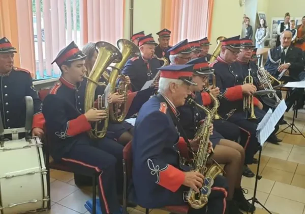 Orkiestra dęta w historycznych mundurach z czerwonymi mankietami i kapeluszami gra na instrumentach dętych w sali przed widownią.