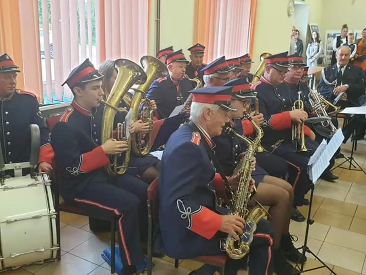 Orkiestra dęta w historycznych mundurach z czerwonymi mankietami i kapeluszami gra na instrumentach dętych w sali przed widownią.