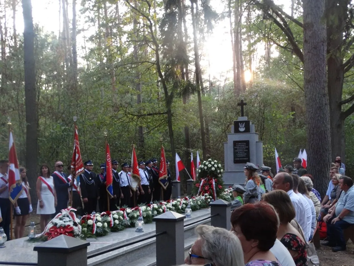 Uroczystość upamiętniająca wybuch II wojny światowej w Powiecie Płockim: uczestnicy w mundurach i strojach cywilnych stoją przed pomnikiem, otoczeni flagami i wieńcami.