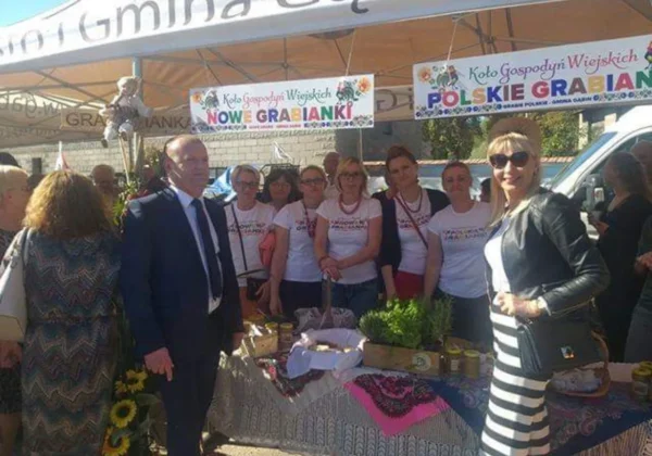 Grupa ludzi stojąca przy stoisku z regionalnymi przysmakami podczas Święta plonów w Dobrzykowie, pod bannerami Koła Gospodyń Wiejskich.