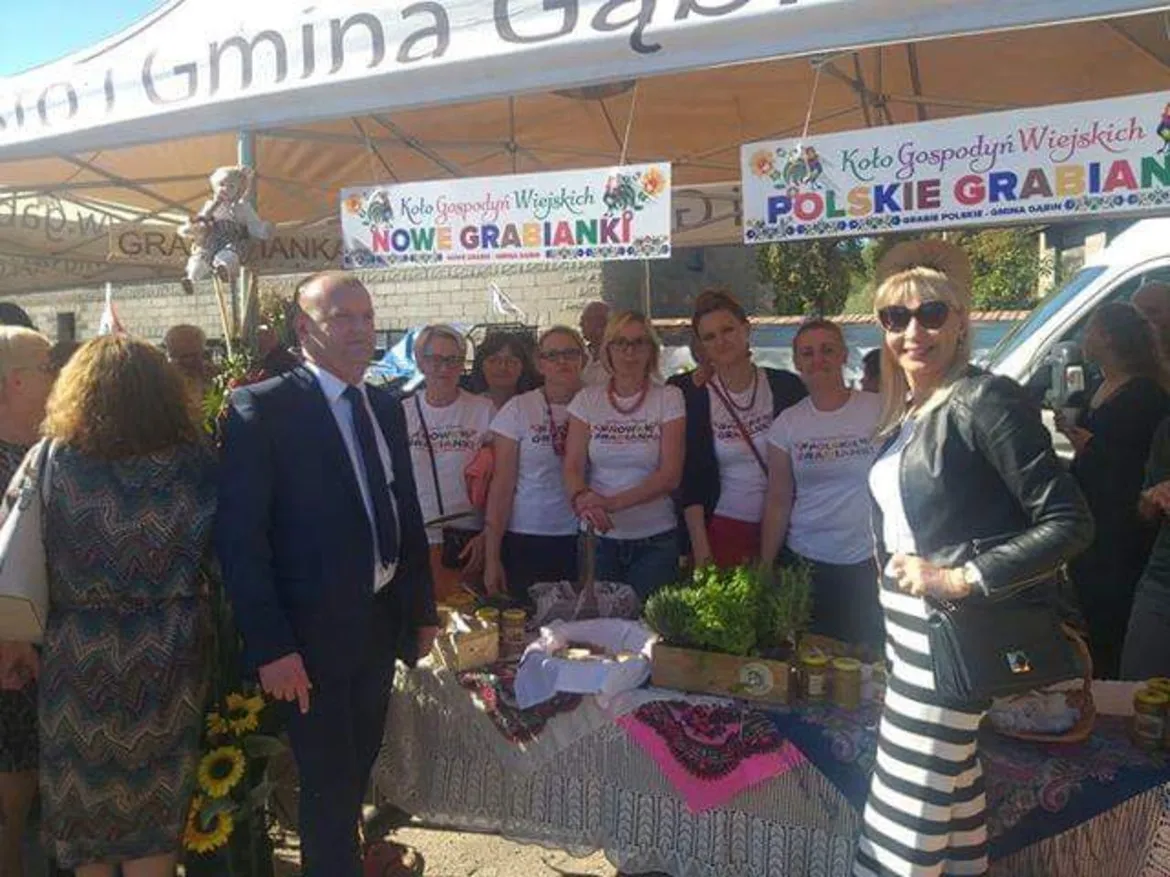 Grupa ludzi stojąca przy stoisku z regionalnymi przysmakami podczas Święta plonów w Dobrzykowie, pod bannerami Koła Gospodyń Wiejskich.