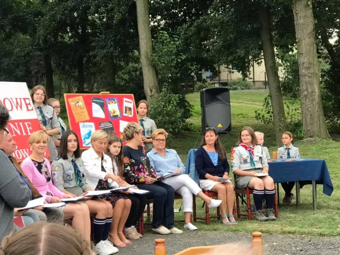 Grupa osób, w tym młodzież w mundurach harcerskich, siedzi w parku podczas ósmej edycji Narodowego Czytania.