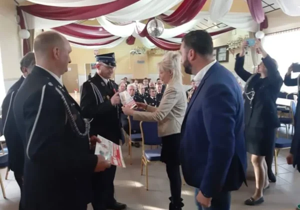 Uroczystość wręczania upominków dla Ochotniczych Straży Pożarnych: kobieta przekazuje prezent mężczyźnie w mundurze strażackim wśród zgromadzonych gości i fotografujących.