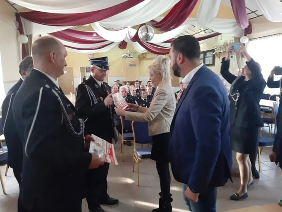 Uroczystość wręczania upominków dla Ochotniczych Straży Pożarnych: kobieta przekazuje prezent mężczyźnie w mundurze strażackim wśród zgromadzonych gości i fotografujących.