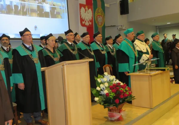 Grupa profesorów w czarnych togach z zielonymi wstęgami i złotymi łańcuchami stoi na scenie podczas inauguracji roku akademickiego na Politechnice Warszawskiej Filii w Płocku.