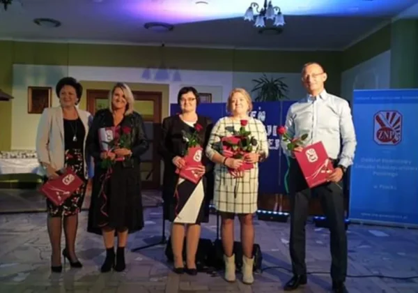 Pięć osób stoi na scenie podczas Powiatowego Dnia Edukacji w Soczewce, trzymając róże i czerwone teczki.