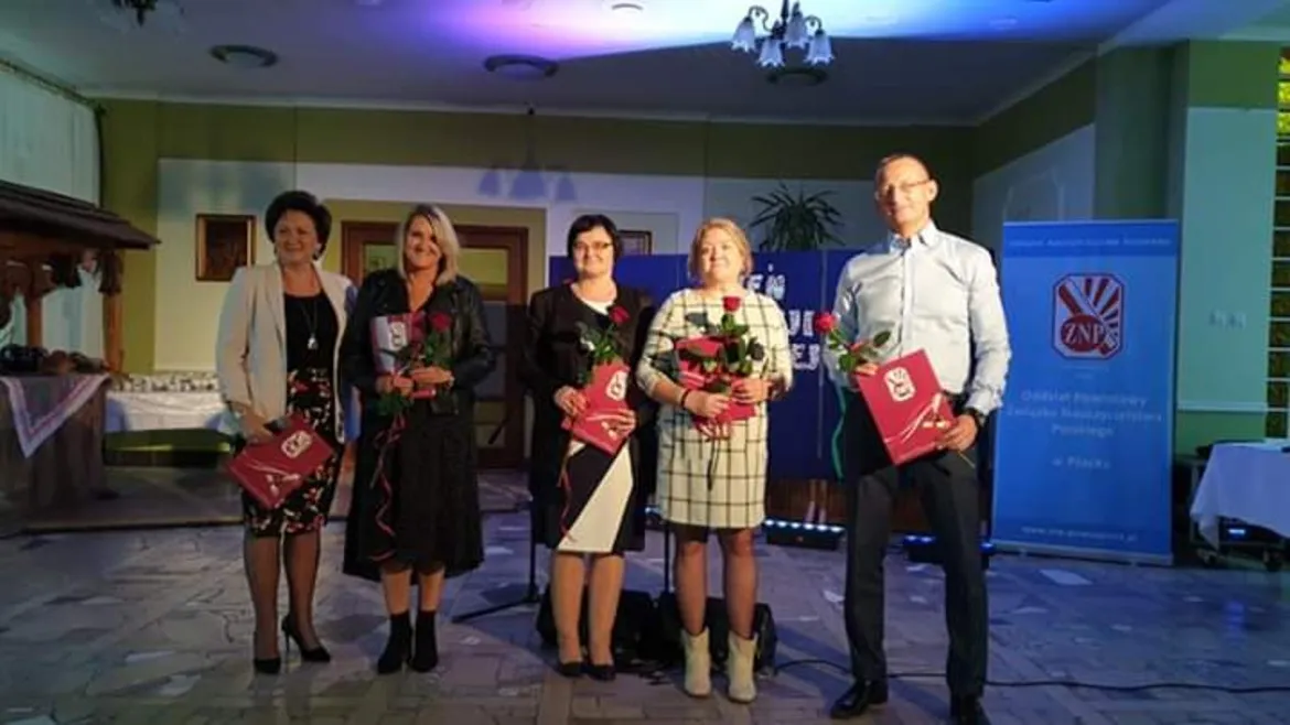 Pięć osób stoi na scenie podczas Powiatowego Dnia Edukacji w Soczewce, trzymając róże i czerwone teczki.