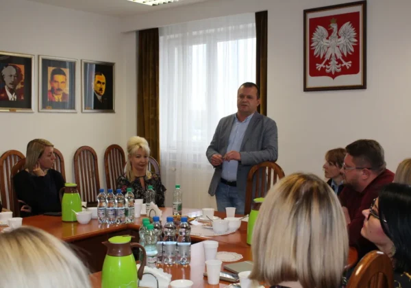 Spotkanie dotyczące ekonomii społecznej w regionie płockim: grupa osób zasiada przy stole w sali konferencyjnej, stojący mężczyzna wygłasza przemówienie, w tle widoczne godło Polski i trzy portrety.