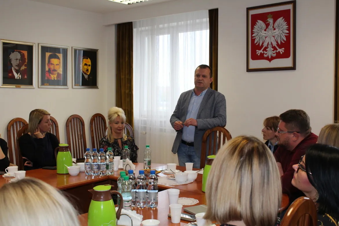 Spotkanie dotyczące ekonomii społecznej w regionie płockim: grupa osób zasiada przy stole w sali konferencyjnej, stojący mężczyzna wygłasza przemówienie, w tle widoczne godło Polski i trzy portrety.