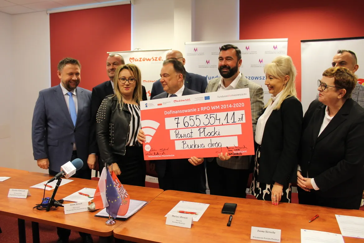 Grupa urzędników na konferencji prezentuje duży czerwono-biały czek z dofinansowaniem w wysokości 7 665 354,11 zł na budowę drogi w powiecie płockim.