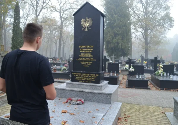 Młody mężczyzna stojący plecami do kamery na cmentarzu, przed wysokim granitowym nagrobkiem z orłem i złotymi inskrypcjami, w otoczeniu grobów, kwiatów i jesiennego klimatu.