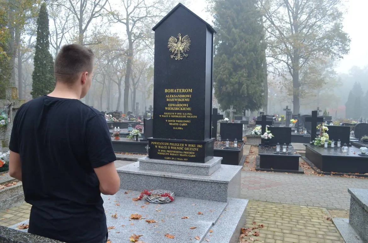Młody mężczyzna stojący plecami do kamery na cmentarzu, przed wysokim granitowym nagrobkiem z orłem i złotymi inskrypcjami, w otoczeniu grobów, kwiatów i jesiennego klimatu.