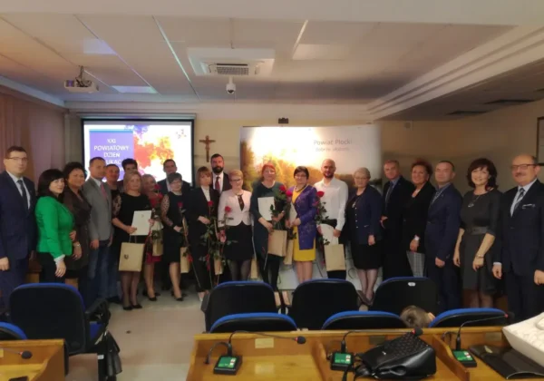 Grupa osób w formalnych ubraniach stoi razem w sali konferencyjnej podczas XXI Powiatowego Dnia Edukacji Narodowej, część z nich trzyma kwiaty i dyplomy.