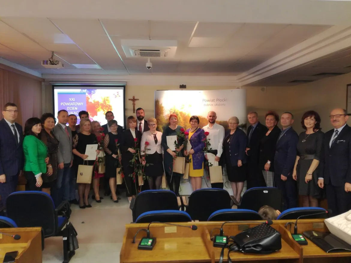 Grupa osób w formalnych ubraniach stoi razem w sali konferencyjnej podczas XXI Powiatowego Dnia Edukacji Narodowej, część z nich trzyma kwiaty i dyplomy.