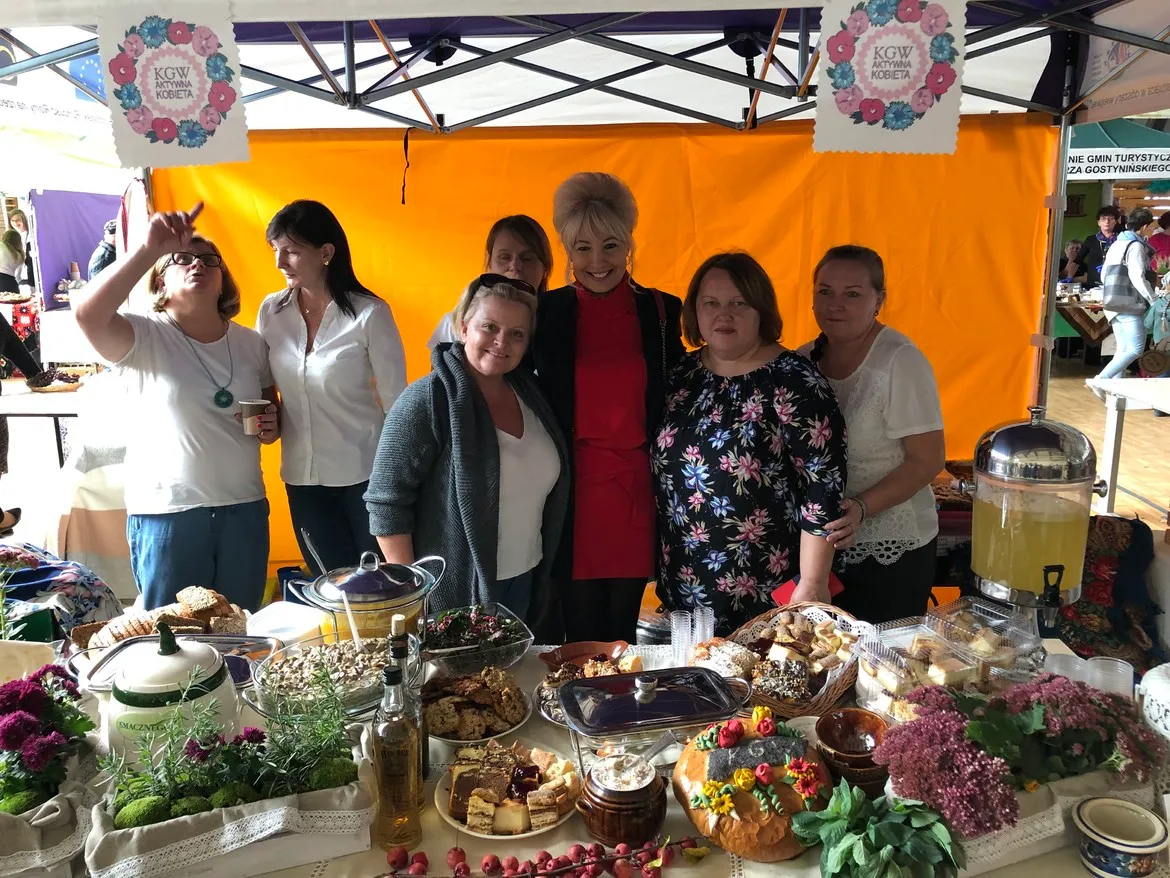 Grupa kobiet stojących za stołem zastawionym różnorodnymi potrawami i napojami pod żółtym namiotem podczas niedzielnego, barwnego wydarzenia w Łącku.