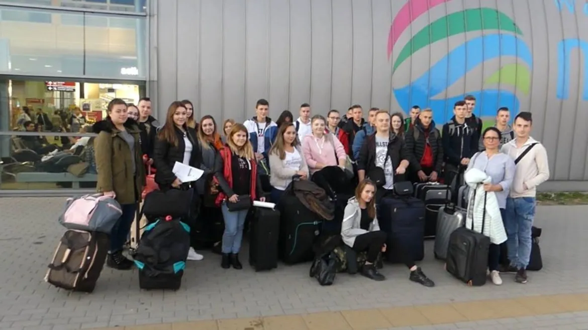 Grupa 26 uczniów Zespołu Szkół im. Jana Śniadeckiego w Wyszogrodzie stoi z walizkami przed nowoczesnym budynkiem z kolorowym muralem, przygotowując się do wyjazdu w ramach ERASMUS+ vol 1.