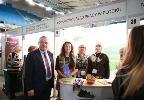 Grupa osób stoi przy stoisku Powiatowego Urzędu Pracy w Płocku na Płockich Targach Aktywności i Przedsiębiorczości.