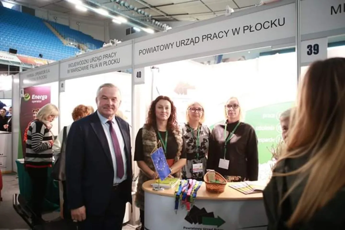 Grupa osób stoi przy stoisku Powiatowego Urzędu Pracy w Płocku na Płockich Targach Aktywności i Przedsiębiorczości.
