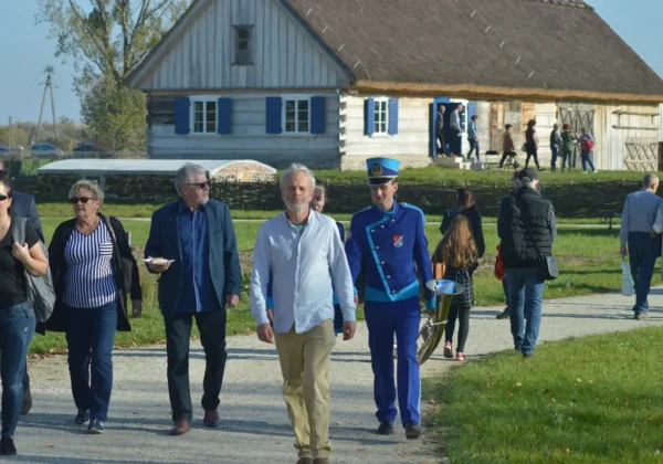Grupa ludzi spaceruje po Skansenie Osadnictwa Nadwiślańskiego w Wiączeminie, mijając drewniany dom ze strzechą w tle.