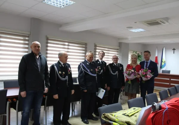 Grupa strażaków w czarnych mundurach z odznaczeniami stoi w sali konferencyjnej gminy Słupno, obok dwie osoby z bukietami, a na stole widoczne elementy sprzętu i umundurowania strażackiego.