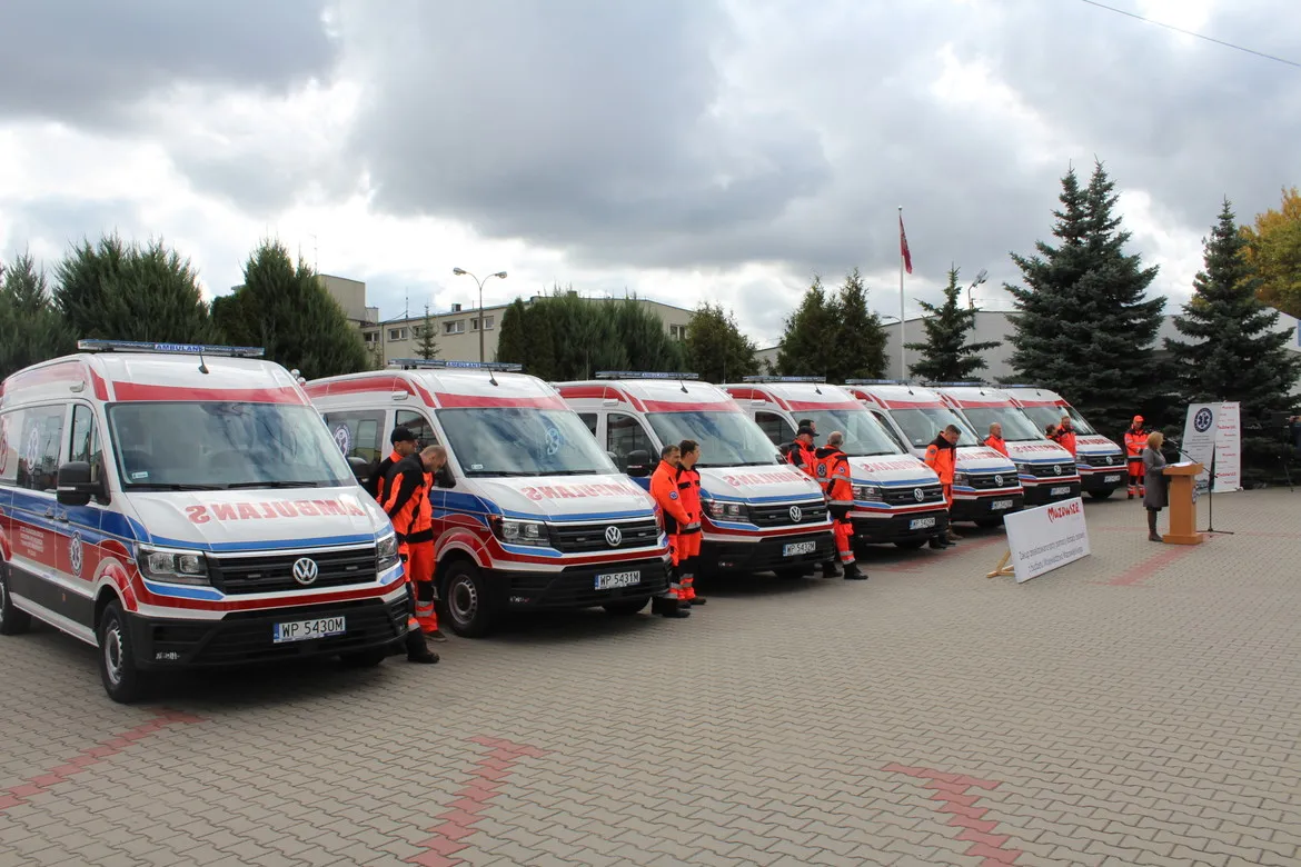 Na placu stoi linia nowych karetek pogotowia w biało‑czerwonych barwach, otoczonych ratownikami w pomarańczowych uniformach podczas uroczystej prezentacji.
