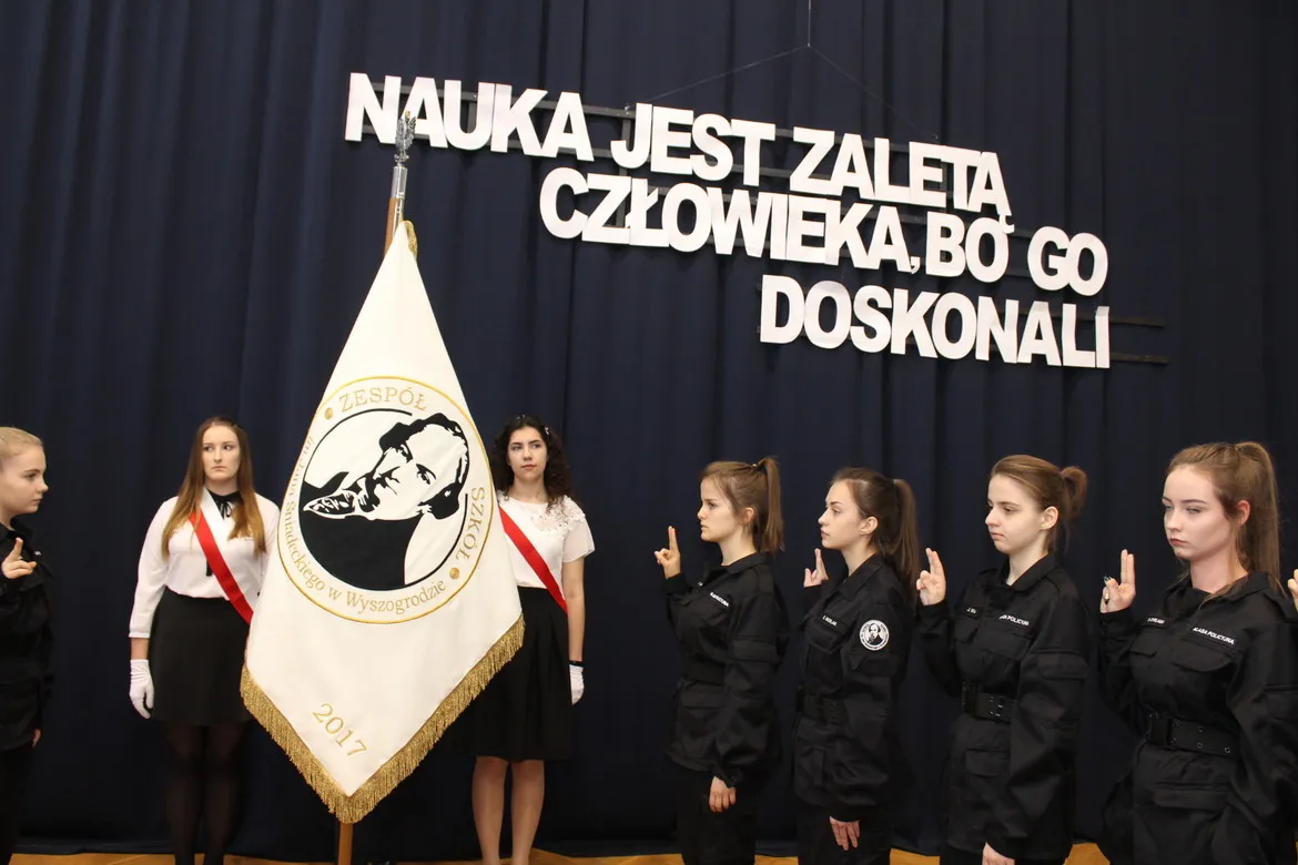 Grupa uczniów klas pierwszych w szkolnych mundurach składa przysięgę podczas obchodów Święta Patrona Szkoły w Zespole Szkół im. Jana Śniadeckiego w Wyszogrodzie, przy sztandarze szkoły.
