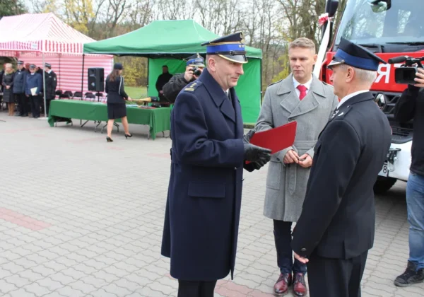 Podczas uroczystości przekazania średniego samochodu ratowniczo-gaśniczego dla Słupna dwie osoby w mundurach wręczają dokumenty przy czerwonym samochodzie strażackim, w tle stoją namioty i zgromadzeni goście.