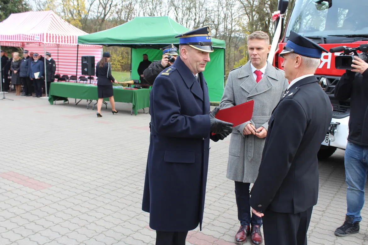 Podczas uroczystości przekazania średniego samochodu ratowniczo-gaśniczego dla Słupna dwie osoby w mundurach wręczają dokumenty przy czerwonym samochodzie strażackim, w tle stoją namioty i zgromadzeni goście.