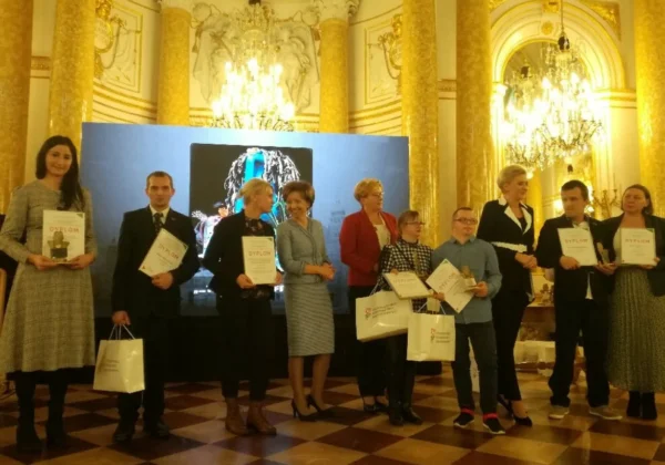 Grupa osób w eleganckich strojach stoi na scenie w bogato zdobionej sali Zamku Królewskiego, trzymając dyplomy i torby z upominkami.