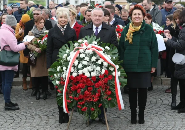 Grupa ludzi stoi na placu podczas obchodów 101. rocznicy Odzyskania Niepodległości, otaczając biało-czerwony wieńiec z kwiatów.