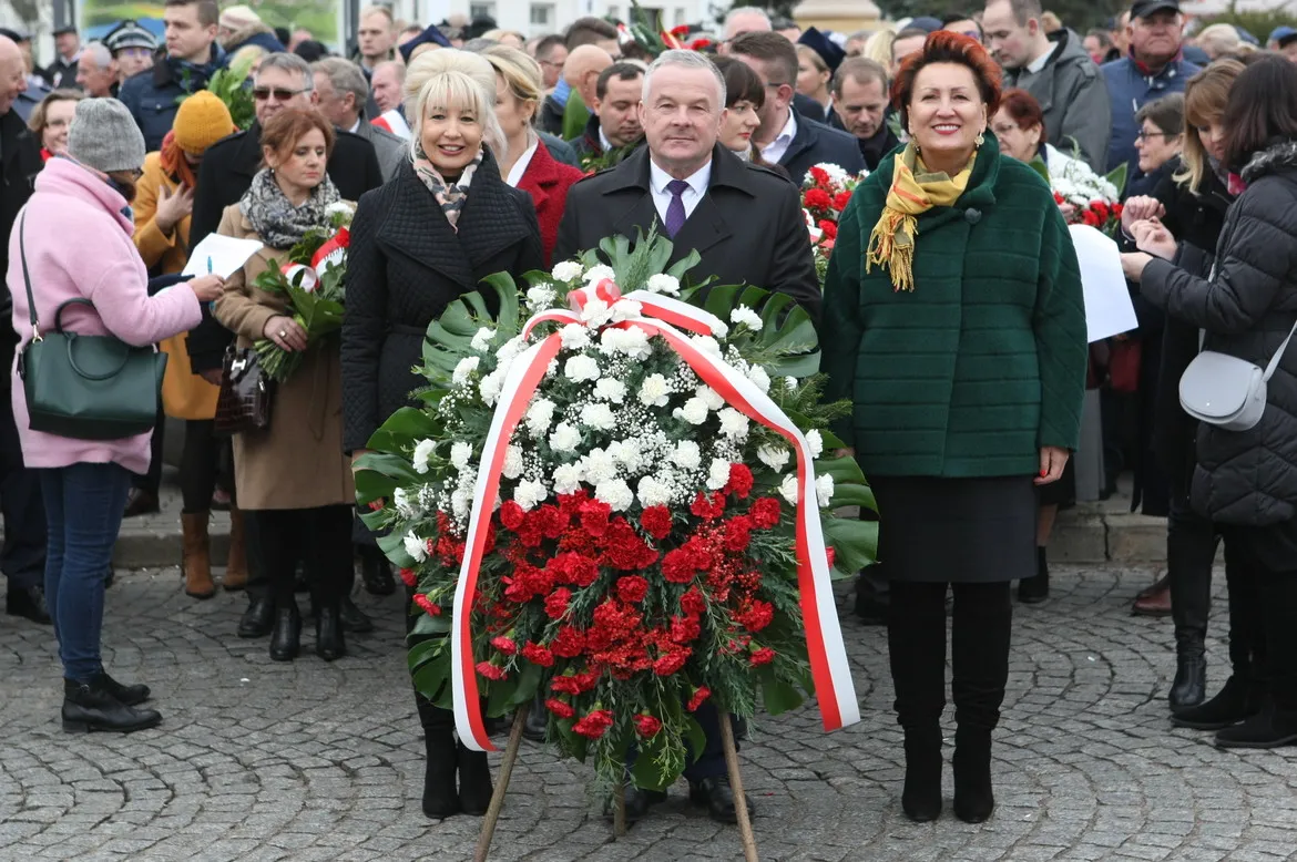 Grupa ludzi stoi na placu podczas obchodów 101. rocznicy Odzyskania Niepodległości, otaczając biało-czerwony wieńiec z kwiatów.