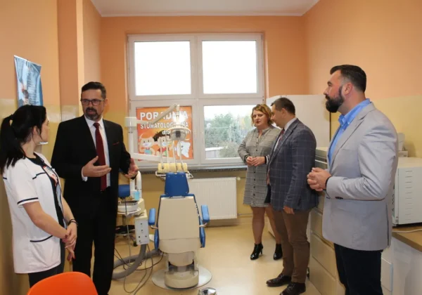 Pięć osób stoi w nowoczesnym gabinecie stomatologicznym w Powiecie Płockim, obok fotela dentystycznego i sprzętu, przy oknie z plakatem „Poradnia Stomatologiczna” na ścianie.