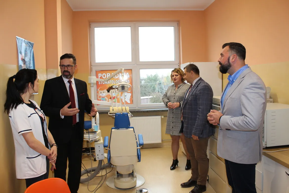 Pięć osób stoi w nowoczesnym gabinecie stomatologicznym w Powiecie Płockim, obok fotela dentystycznego i sprzętu, przy oknie z plakatem „Poradnia Stomatologiczna” na ścianie.