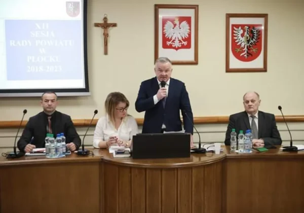 Mężczyzna w garniturze przemawia do mikrofonu podczas XII sesji Rady Powiatu w Płocku w kadencji 2018–2023, na tle sali wiszą krzyż i dwa herby, a przy stole siedzą inni członkowie rady.