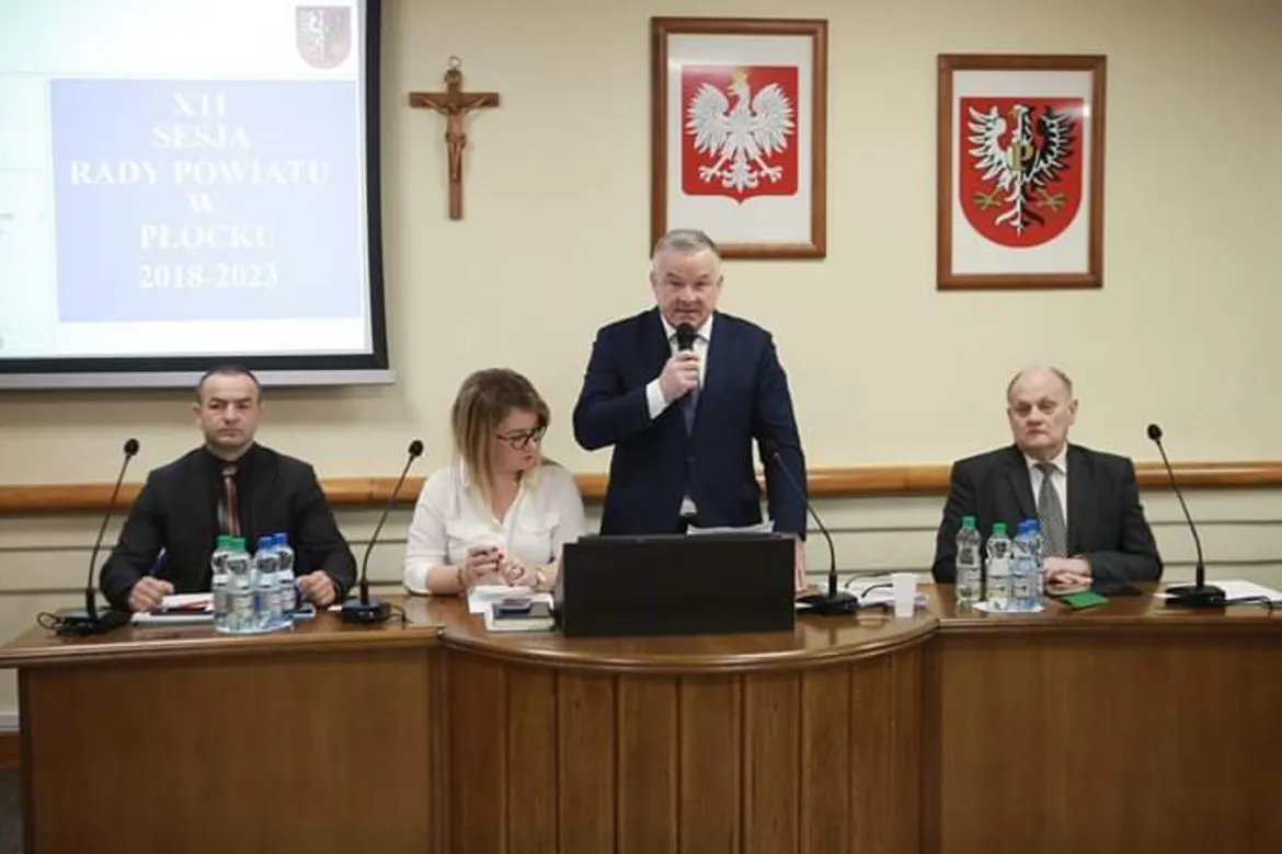 Mężczyzna w garniturze przemawia do mikrofonu podczas XII sesji Rady Powiatu w Płocku w kadencji 2018–2023, na tle sali wiszą krzyż i dwa herby, a przy stole siedzą inni członkowie rady.