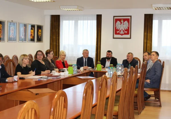 Grupa osób w formalnych ubraniach zasiada przy długim, drewnianym stole podczas posiedzenia Zarządu Powiatu w sali konferencyjnej, a na ścianie wisi godło Polski.