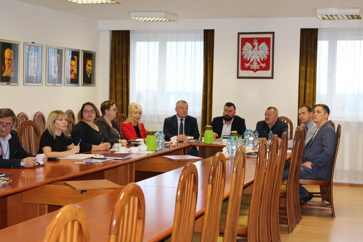 Grupa osób w formalnych ubraniach zasiada przy długim, drewnianym stole podczas posiedzenia Zarządu Powiatu w sali konferencyjnej, a na ścianie wisi godło Polski.