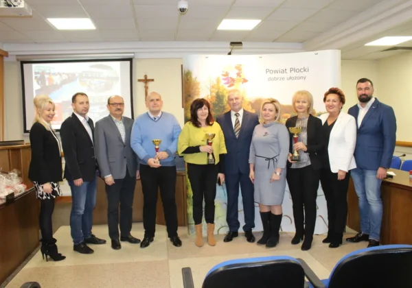 Grupa kilkunastu osób w biznesowych strojach stoi na sali konferencyjnej, część z nich trzyma puchary, podczas podsumowania współzawodnictwa sportowego szkół w powiecie płockim za rok szkolny 2018/2019.