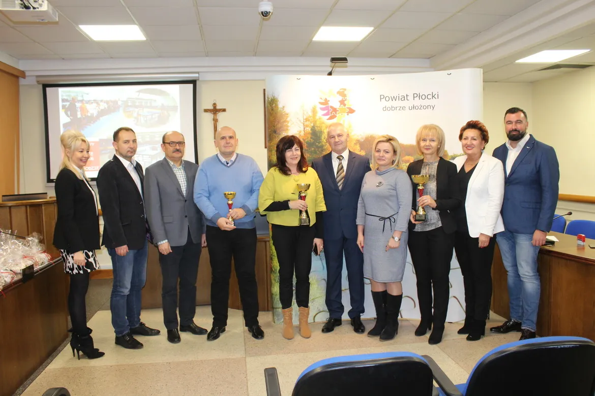 Grupa kilkunastu osób w biznesowych strojach stoi na sali konferencyjnej, część z nich trzyma puchary, podczas podsumowania współzawodnictwa sportowego szkół w powiecie płockim za rok szkolny 2018/2019.