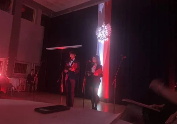 Dwoje uczniów stoi na scenie i przemawia do mikrofonów podczas szkolnych obchodów 101. rocznicy odzyskania niepodległości, a w tle widoczna jest biało-czerwona flaga z orłem.