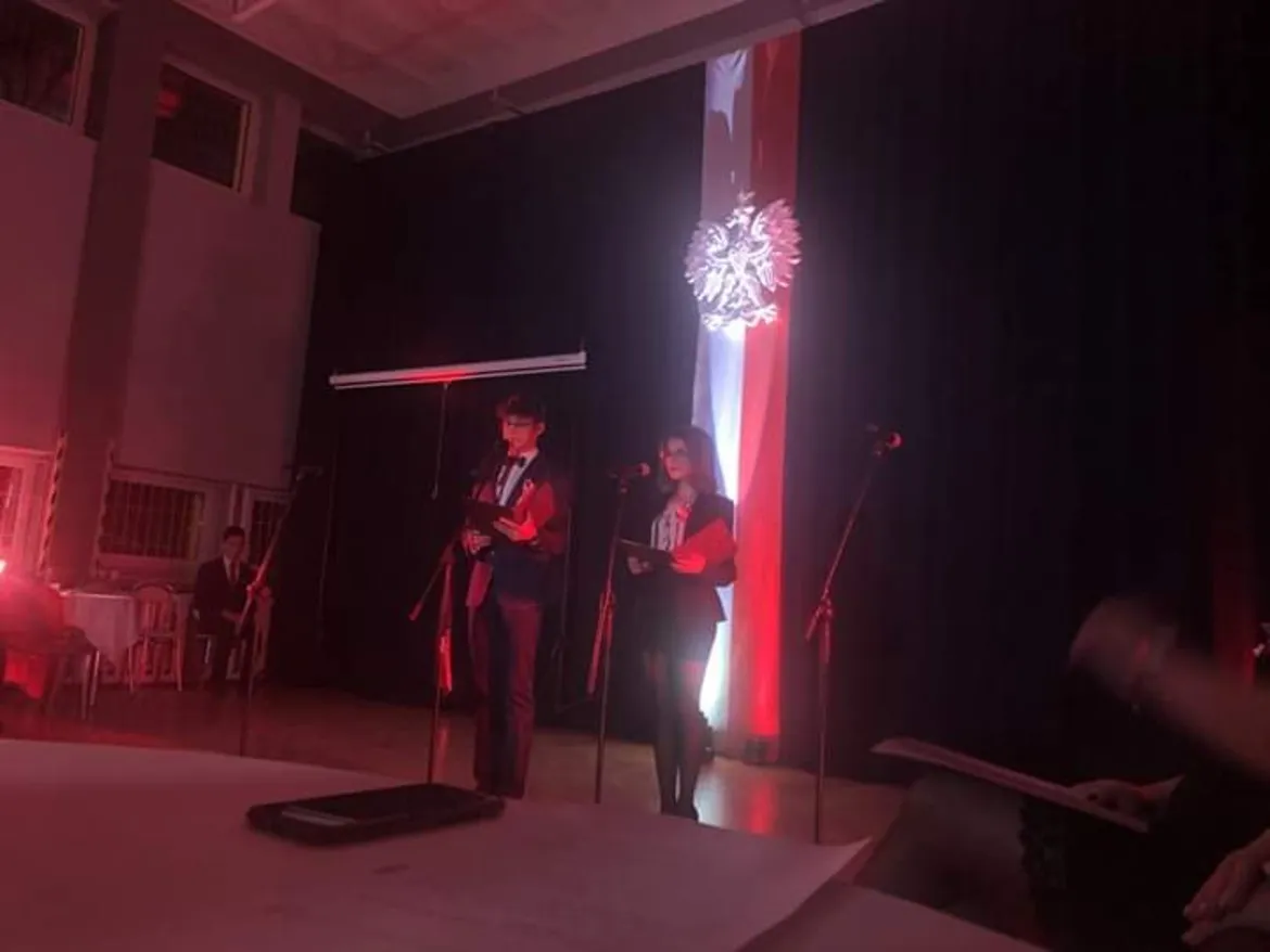 Dwoje uczniów stoi na scenie i przemawia do mikrofonów podczas szkolnych obchodów 101. rocznicy odzyskania niepodległości, a w tle widoczna jest biało-czerwona flaga z orłem.