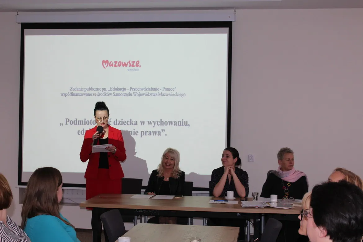 Prelegentka w czerwonym garniturze przemawia do publiczności z mikrofonem podczas konferencji, na ekranie w tle widnieje tytuł „Podmiotowość dziecka w wychowaniu, edukacji i systemie prawa”.