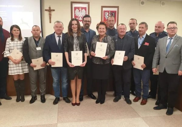 Grupa kilkunastu osób w formalnych strojach stoi w sali konferencyjnej, każdy trzyma certyfikat, na tle widoczne polskie godła i krzyż — wydarzenie mające na celu poprawę bezpieczeństwa na drogach.