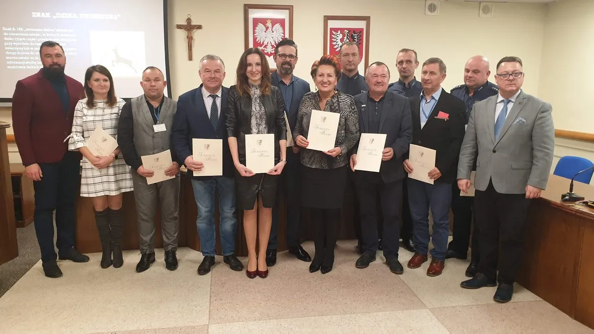 Grupa kilkunastu osób w formalnych strojach stoi w sali konferencyjnej, każdy trzyma certyfikat, na tle widoczne polskie godła i krzyż — wydarzenie mające na celu poprawę bezpieczeństwa na drogach.