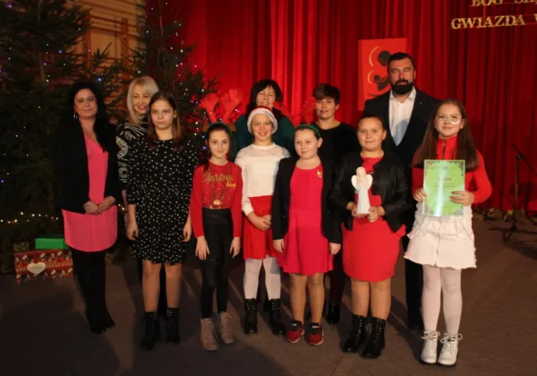 Grupa uczniów i nauczycieli stoi na scenie w Zespole Szkół w Gąbinie podczas bożonarodzeniowego występu „Bóg się rodzi, gwiazda wschodzi”, w tle choinki i czerwone zasłony.