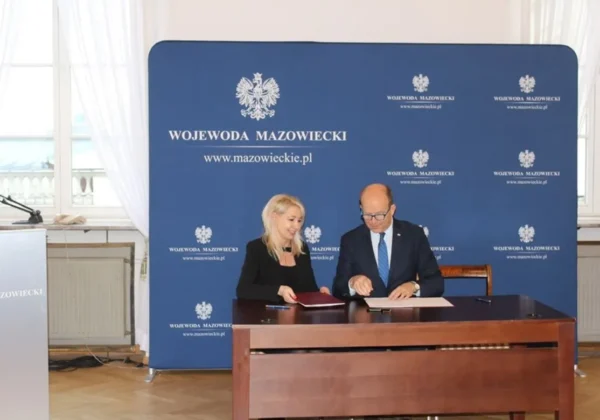 Dwie osoby siedzą przy stole i podpisują dokumenty podczas podpisywania umów o dofinansowanie ze środków Funduszu Dróg Samorządowych, na tle niebieskiej ścianki z napisem Wojewoda Mazowiecki.