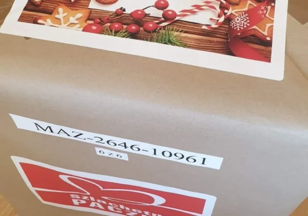 Pudełko paczki z ozdobną kartką bożonarodzeniową z motywem pierników i cukierków oraz napisem „Wesołych Świąt”, leżące na drewnianej podłodze.