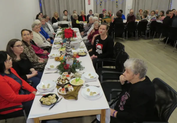 Grupa seniorów i opiekunów siedzi przy długim, świątecznie udekorowanym stole podczas Wigilii Płockiego Stowarzyszenia Pomocy Osobom z Chorobą Alzheimera.