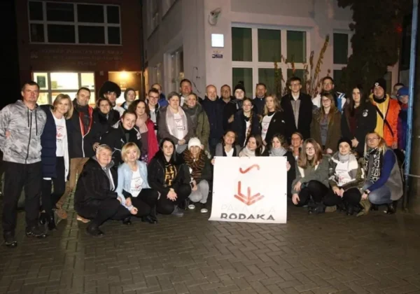 Grupa ludzi stojących na chodniku wieczorem, trzymająca baner z napisem „Paka dla Rodaka”.